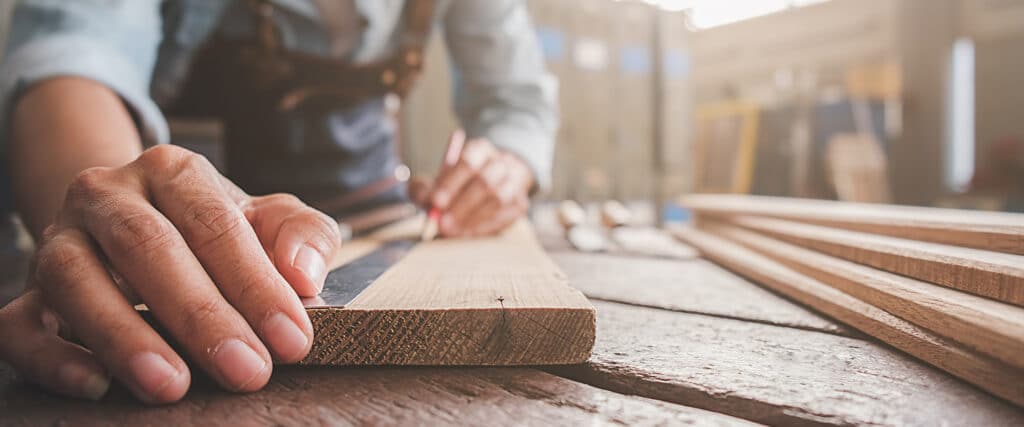 Menuisier Expert: Mesure Précise pour vos Projets Bois Menuisier mesurant une planche de bois avec une règle et un crayon sur un établi d'atelier. Mains concentrées sur le travail du bois.