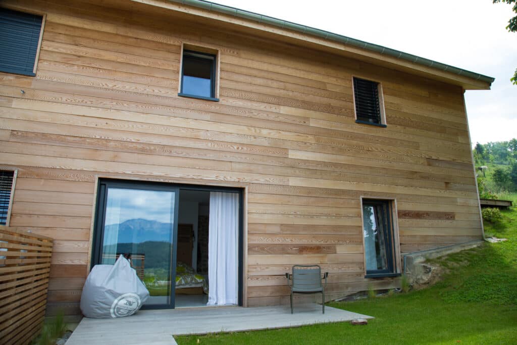 Maison en Bois Moderne: Design & Vue Montagne Panoramique Maison moderne en bois clair avec grandes baies vitrées et terrasse en bois. Paysage montagneux en arrière-plan. Sac pouf North Sails.