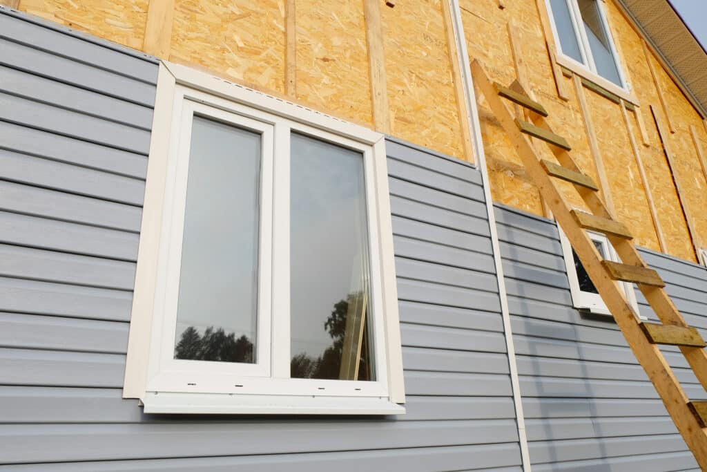 Façade de maison en construction avec revêtement vinyle gris et bardage OSB apparent, une fenêtre blanche installée et une échelle en bois.