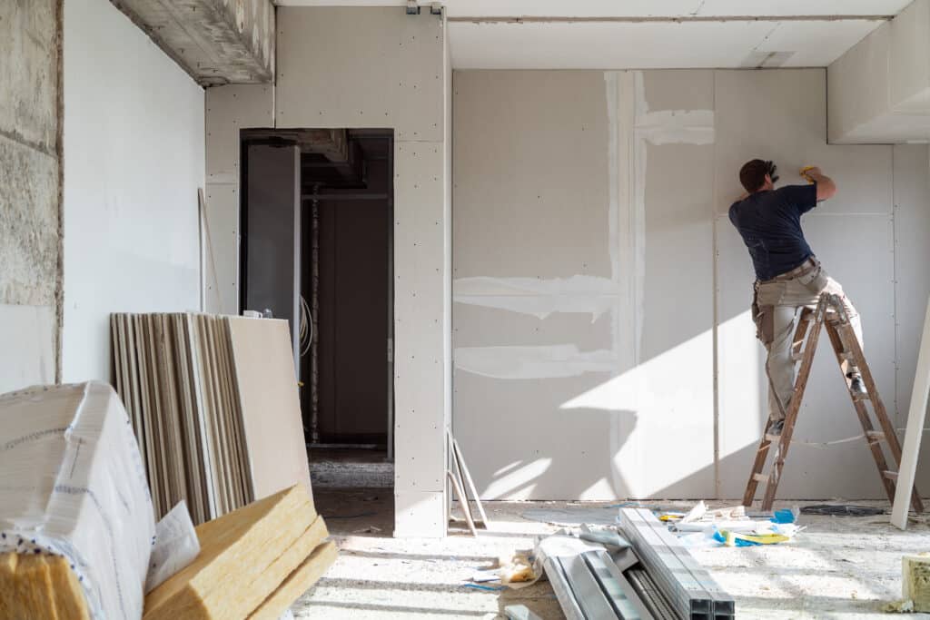 Ouvrier sur échelle installant des plaques de plâtre sur un mur lors de travaux de rénovation intérieure, avec matériaux de construction.