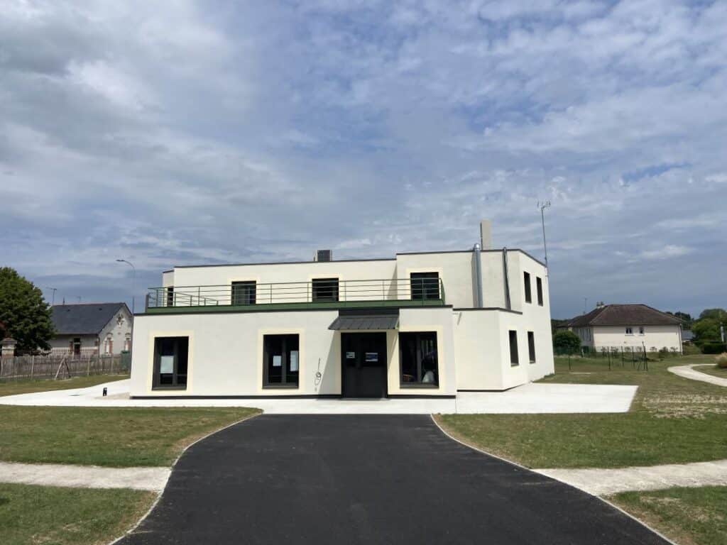 Bâtiment moderne blanc style paquebot avec balcon, entouré d'herbe et d'un accès asphalté neuf sous ciel nuageux.