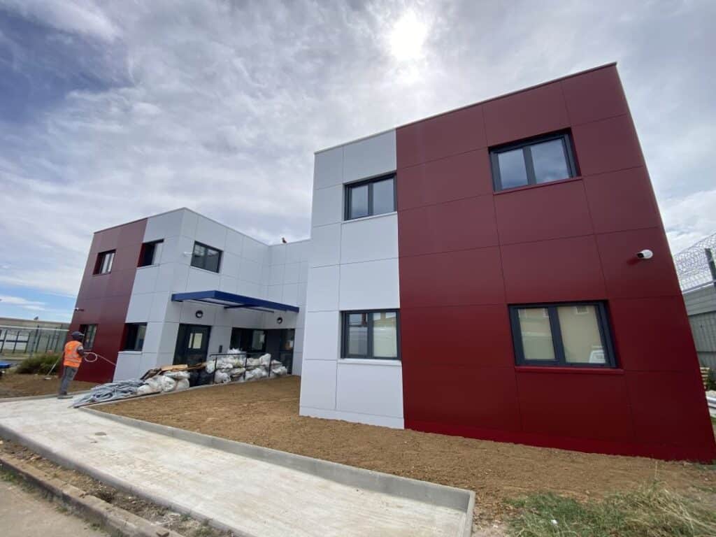 Construction d'un bâtiment moderne bicolore rouge bordeaux et blanc avec ouvrier en gilet orange devant, ciel nuageux.