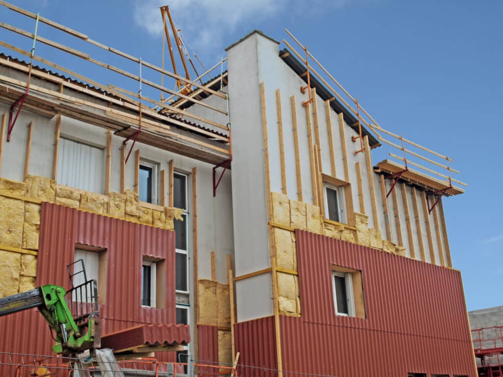Rénovation Façade : Isolation et Bardage en Chantier Chantier de rénovation énergétique : pose d'isolant laine minérale et de bardage ondulé rouge sur façade, avec échafaudage.